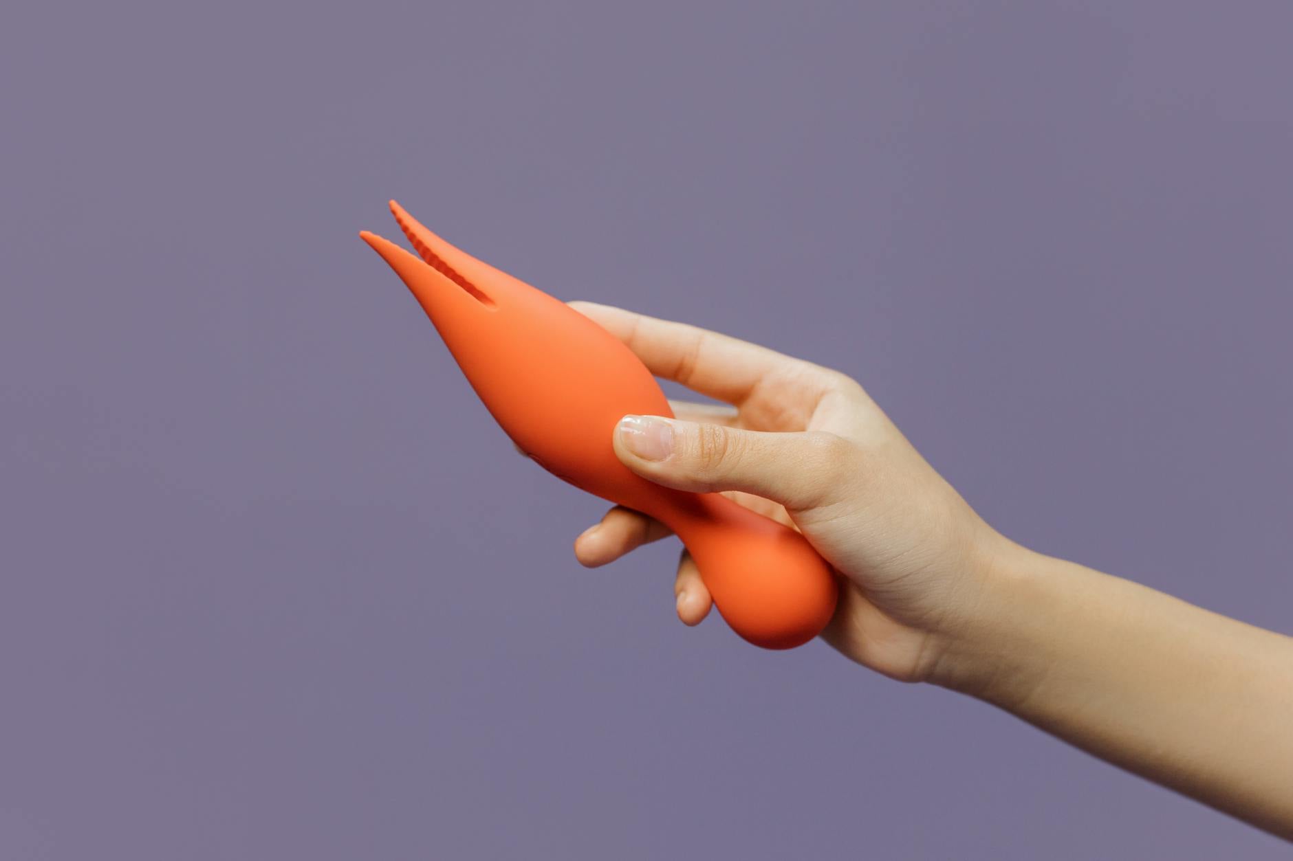 Mãos segurando vibradores de silicone em tons pastel contra fundo roxo mínimalista, demonstrando conforto e autossuficiência.